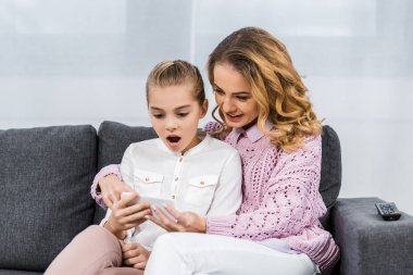 kanepede oturan ve sürpriz kızı smartphone ekran oturma odasında bakarak kucaklayan güzel kadın