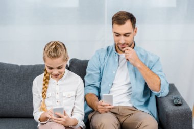 gülümseyen baba ve kızı koltukta oturan ve akıllı telefonlar oturma odasında kullanarak