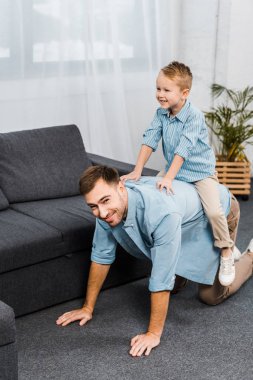 smiling boy sitting on back of father standing on all fours and looking at camera in living room clipart