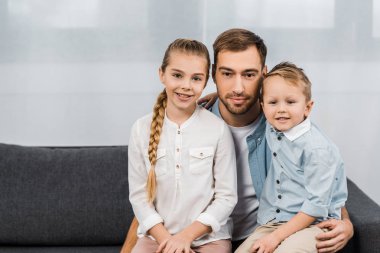 smiling father with cute children sitting on sofa and looking at camera in apartment clipart