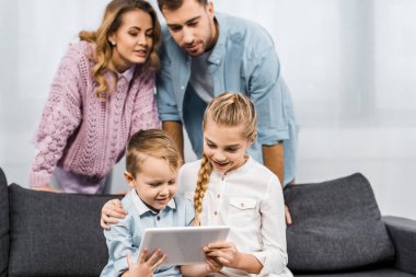 ayakta ve kanepede oturan ve dijital tablet oturma odasında kullanma gülümseyen kardeşler bakarak anne