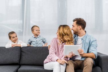 parents sitting on sofa, holding digital tablet and looking at children in living room clipart