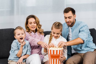 mutlu aile koltukta oturan, tv izlerken ve oturma odasında çizgili kova patlamış mısır yemek