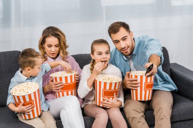 kanepede oturan ve aile çizgili kova tutarak ve dairede patlamış mısır yemek ile uzaktan kumanda kanalları değiştirme babası gülümseyen