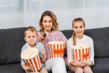 smiling mother with children sitting on sofa, holding striped popcorn buckets and changing channels by remote controller in living room clipart