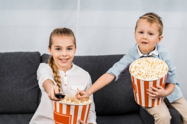 smiling siblings sitting on sofa, holding striped popcorn buckets and changing channels by remote controller clipart
