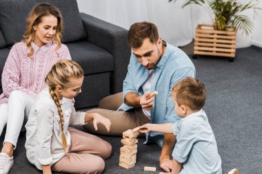 two parents playing blocks wood tower game with children on floor in apartment clipart