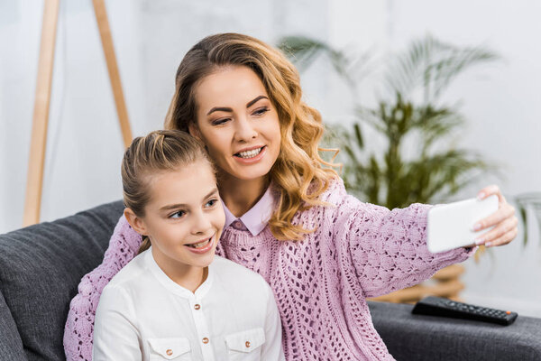happy mother and daughter sitting on sofa and taking selfie in apartment