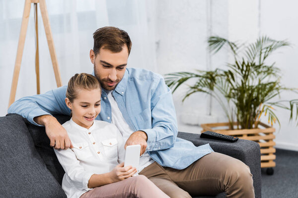 smiling daughter sitting on sofa and father pointing with finger at screen in apartment