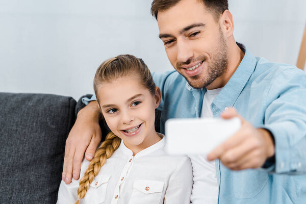 cheerful father sitting on sofa, embracing daughter and taking selfie in apartment