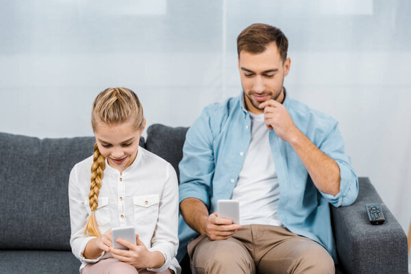 smiling father and daughter sitting on sofa and using smartphones in living room