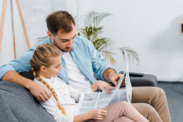 father and daughter sitting on sofa and reading newspaper in living room