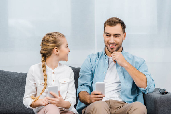 smiling father and daughter sitting on sofa, holding smartphones and looking at each other in living room