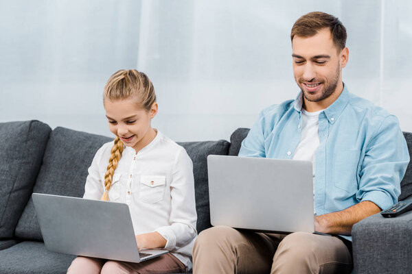 smiling daughter and father sitting on sofa and typing on laptop keyboards in living room