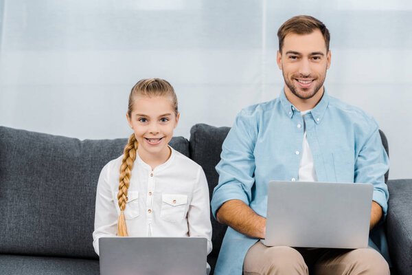 smiling daughter and father sitting on sofa, holding laptops and looking at camera in living room