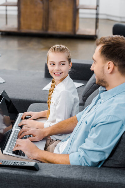 smiling father and daughter sitting on sofa, typing on laptop keyboards and looking at each other