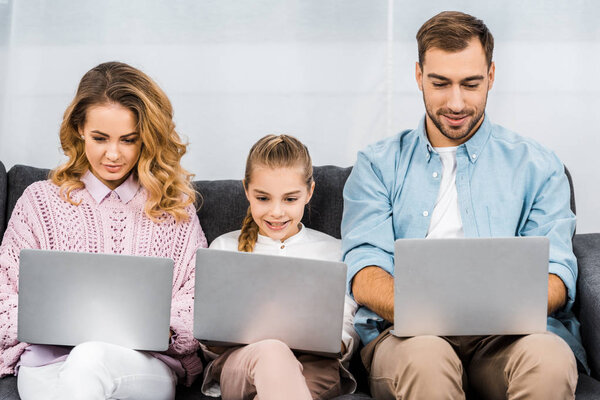 cute girl with two parents using laptops and sitting on sofa in living room