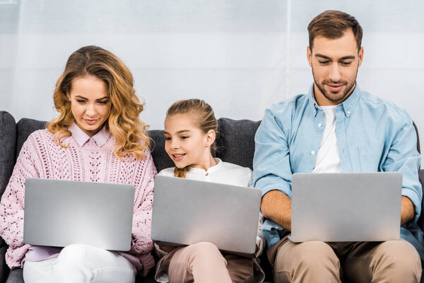 cute girl with two parents sitting on sofa and using laptops in apartment