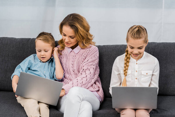 pretty woman with son and daughter sitting on sofa and using laptops in apartment