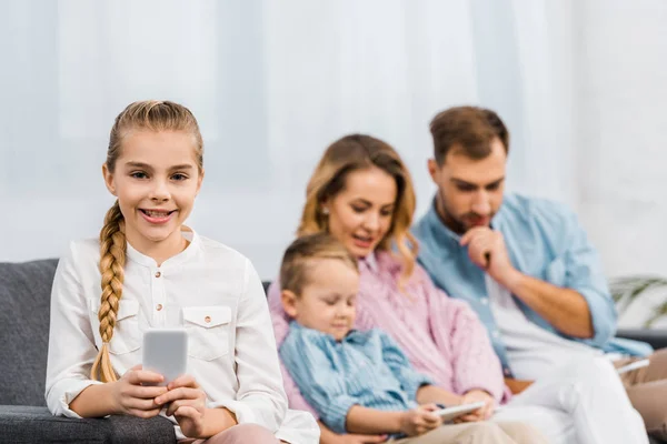 Smiling Girl Sitting Sofa Holding Smartphone Looking Camera Family Background — Free Stock Photo