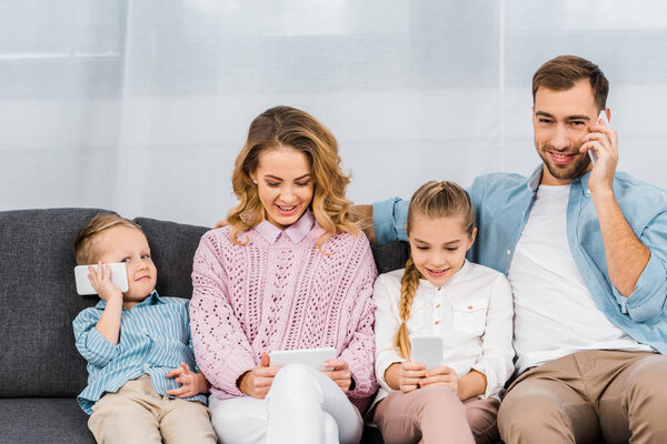 smiling father sitting on sofa and talking on phone while mother and children using smartphones in apartment