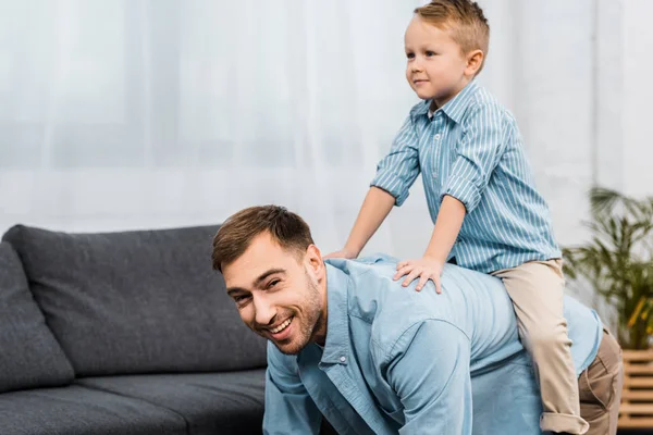 father standing on all fours and looking at camera while cute boy sitting on back in apartment