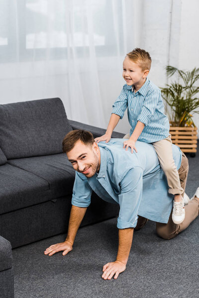 smiling boy sitting on back of father standing on all fours and looking at camera in living room
