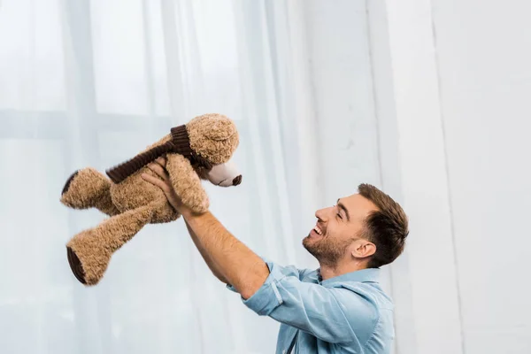 smiling man holding teddy bear in raising hands in living room