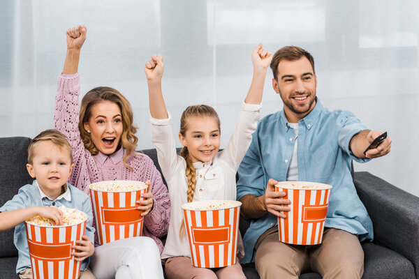 smiling father sitting on sofa and changing channels by remote controller with wife and daughter raising hands and son eating popcorn in apartment
