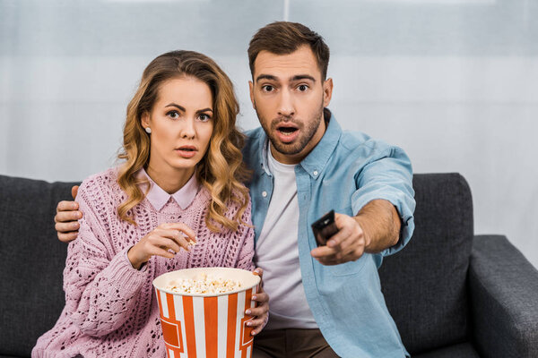 open-eyed man sitting on sofa, holding remote controller and embracing surprised wife with striped popcorn bucket