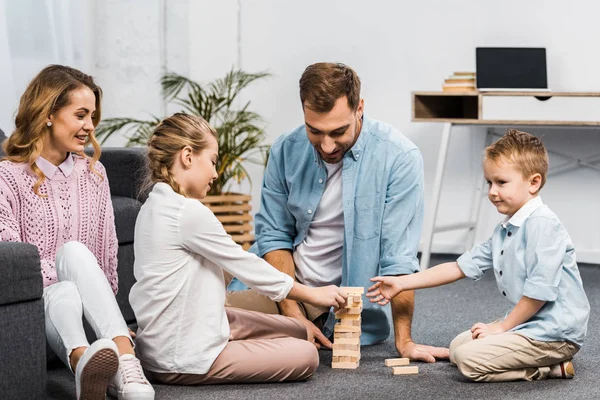 two parents playing blocks wood tower game with daughter and son on floor in living room