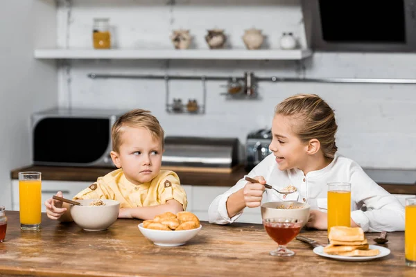 Smiling Girl Holding Spoon Porridge Looking Upset Little Brother Table — Stock Photo, Image