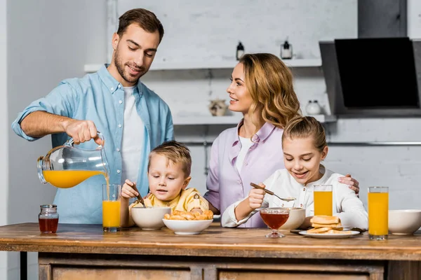 微笑的妇女看着丈夫倒在玻璃橙汁 而孩子吃燕麦片 — 图库照片