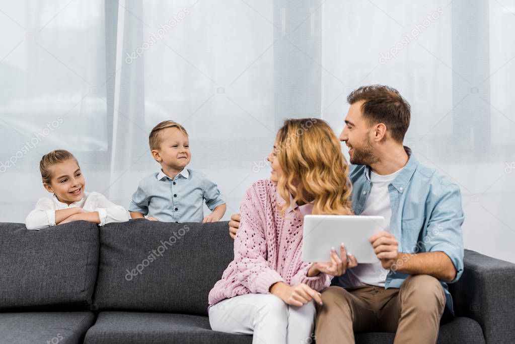 parents sitting on sofa, holding digital tablet and looking at children in living room