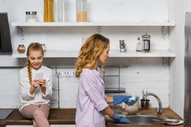 attractive woman in rubber gloves washing up while daughter sitting on table and using smartphone in kitchen clipart