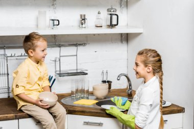 cute girl in rubber gloves washing dishes and looking at smiling boy sitting on table and holding bowl in kitchen clipart