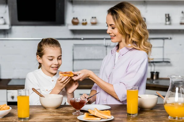 smiling woman giving toast with jam to cute daughter at table in kitchen
