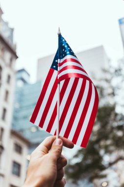 cropped shot of man holding american flag in hand with blurred new york city street on background clipart