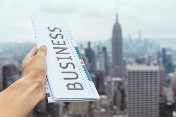частичный взгляд человека, держащего деловую газету с размытым видом на город Нью-Йорк на заднем плане

