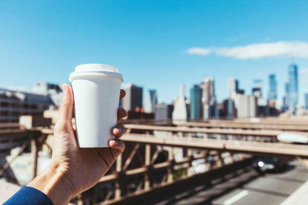 partial view of man holding disposable cup of coffee with blurred new york city on background