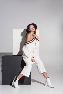 çekici Afrikalı-Amerikalı kadın moda beyaz giysiler ve siyah küp üzerinde oturan ve kameraya beyaz üzerinde seyir şapka