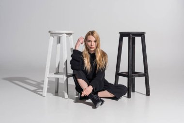 attractive blonde woman in stylish black clothes sitting near chairs and looking at camera on white clipart