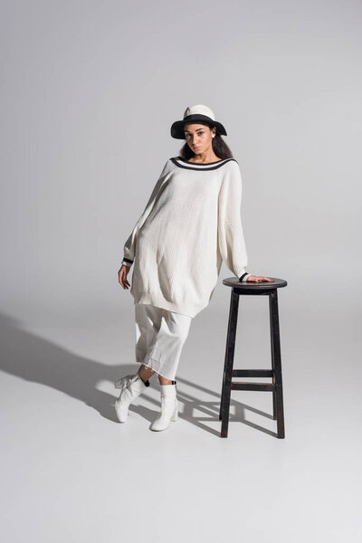 attractive african american girl in stylish white clothes and hat leaning on black chair and looking at camera on white