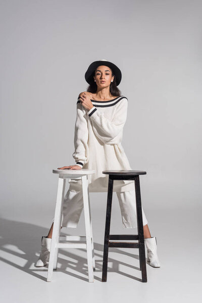 attractive african american girl in stylish white clothes and hat standing near black and white chairs on white