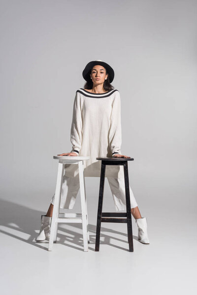 attractive african american girl in fashionable clothes and hat leaning on black and white chairs on white