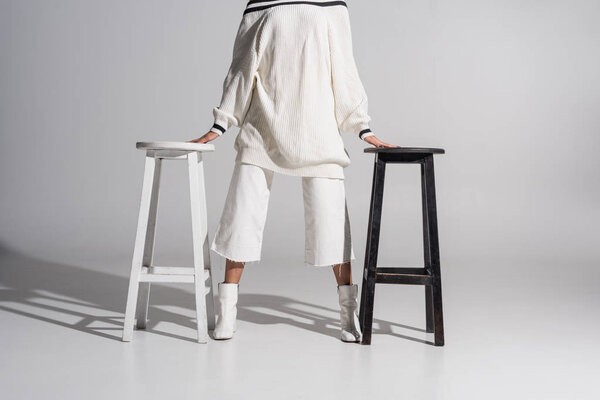 cropped image of girl in fashionable sweater and trousers standing between black and white chairs on white