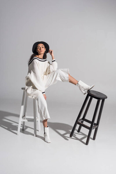 attractive african american girl in stylish white clothes and hat sitting on white chair and looking at camera on white