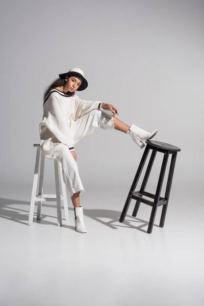 attractive african american model in fashionable white clothes and hat sitting on white chair and looking at camera on white