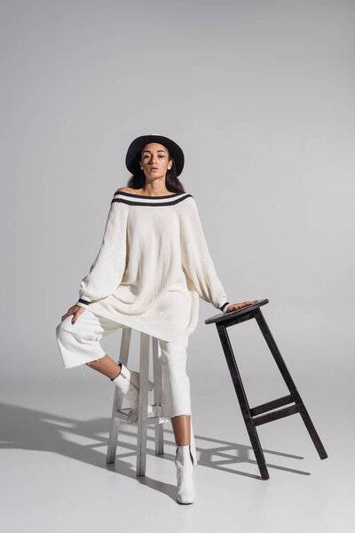 attractive african american girl in stylish white clothes and hat posing on black and white chairs on white
