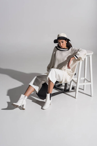 attractive african american girl in stylish white clothes and hat sitting on black and white chairs and looking at camera on white